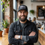 Bearded man in black trucker hat with white slogan patch in casual cafe setting with plants